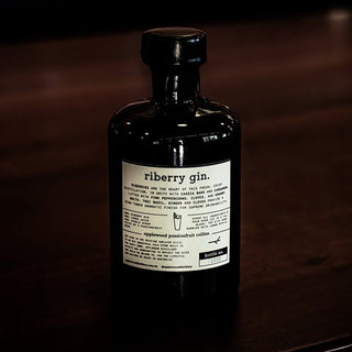 A dark glass bottle of Applewood Riberry Gin, infused with native botanicals, rests on a dark wooden surface. Its white label features the name, tasting notes, and bottling details, while moody lighting enhances its aromatic spice allure.