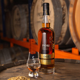 A bottle of Indri Drú Cask Strength Single Malt Indian Whisky stands beside a filled glass on a barrel, with aromas of chocolate and ripe fruit, as blurred grain and whisky barrels appear in the background.