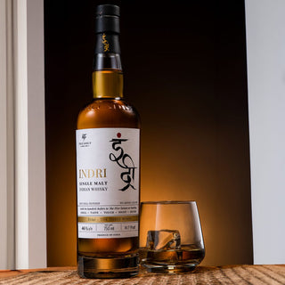 A bottle of Indri Trini Single Malt Indian Whisky stands beside a glass with whisky and ice on a wooden surface, against a dark, softly lit background—evoking notes of warm spice and black tea.
