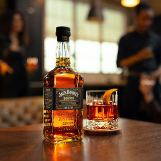 A bottle of Jack Daniel’s Bonded Tennessee Whiskey and a glass garnished with orange peel rest on a wooden table in a cozy, warmly lit bar, as citrus and toasted oak aromas blend amid the lively, blurred crowd in the background.