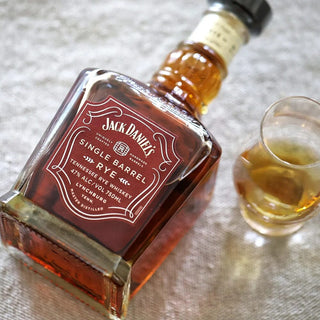 A bottle of Jack Daniel’s Single Barrel 4 Year Old Rye Whiskey rests on its side beside a whiskey-filled glass on a textured light surface, showcasing caramel and toasted oak notes.