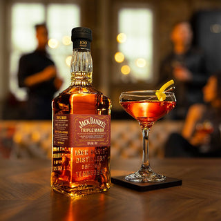 A bottle of Jack Daniel’s Triple Mash Tennessee Whiskey sits on a table next to a cocktail glass with a lemon twist, its rich oak notes suggested. Two blurred figures stand in the warmly lit background.