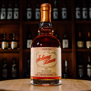 A bottle of Willett Family Estate Johnny Drum Kentucky Bourbon Whiskey sits on a round wooden surface, its Private Stock label displaying rustic, vintage-style typography and notes of oak, caramel, and vanilla. Blurred bottles fill the background shelves.