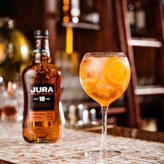 A bottle of Jura 10 Year Old Single Malt Scotch Whisky sits on a bar next to a stemmed glass with an orange cocktail and slice of orange. Notes of vanilla and toasted almonds linger as shelves and bar décor blur in the background.