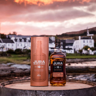 A bottle of Jura 10 Year Old Single Malt Scotch Whisky and its cylindrical box rest on a wooden barrel outdoors, with white houses and a scenic sunset in the background—evoking flavors of dark chocolate and toasted almonds.