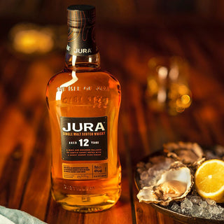 A bottle of Jura 12 Year Old Single Malt Scotch Whisky rests on a wooden table beside oysters on ice and lemon slices, with notes of coffee and honey wafting through the warm, cozy atmosphere.