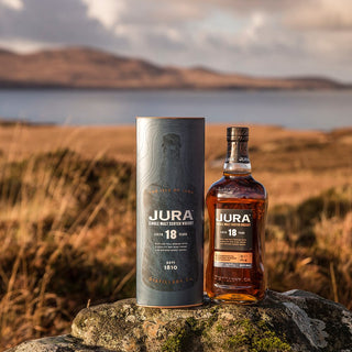 A bottle of Jura 18 Year Old Single Malt Scotch Whisky with its box rests on a rock by calm water amid rugged scenery—the ideal place to enjoy its dark chocolate and black forest fruit notes.