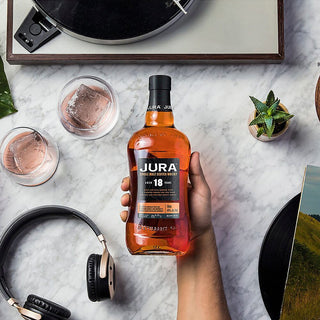 A hand holds a bottle of Jura 18 Year Old Single Malt Scotch Whisky on a marble table, surrounded by two whiskey glasses with ice, a record player, headphones, a plant, and a book—evoking dark chocolate and black forest fruit notes.