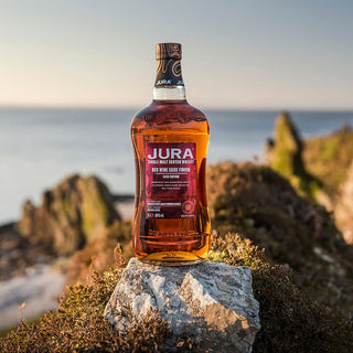 A bottle of Jura Red Wine Cask Finish Single Malt Scotch Whisky stands on a rock with rugged coastline and sea blurred in the background, illuminated by warm natural sunlight, hinting at caramel notes within its rich golden hue.