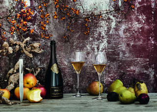 Bottle of champagne with two glasses and fruits on a rustic wooden table against a textured wall.