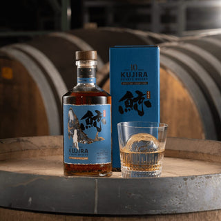A bottle of Kujira Ryukyu 10 Year Old Japanese Whisky, its blue box, and a glass of spiced whisky with ice sit on a wooden barrel in a softly lit distillery, surrounded by large aging barrels in the background.