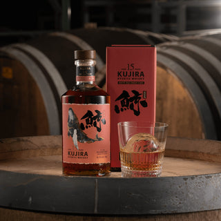 A bottle of Kujira Ryukyu 15 Year Old Japanese Whisky with its red box and a glass rest on a wooden barrel in a dimly lit distillery, tropical fruit notes wafting among rows of aging barrels in the background.