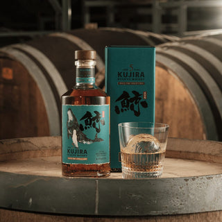 A bottle of Kujira Ryukyu 5 Year Old Japanese Whisky, its box, and a glass with ice rest on a barrel in a dimly lit distillery, where aromas of spice, caramel, and rich oak drift from the wooden barrels in the background.