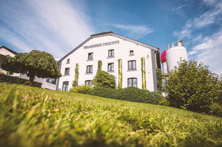 White building with 'Brasserie Chouffe' sign, surrounded by greenery and blue sky.