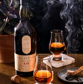 A bottle of Lagavulin 16 Year Old Single Malt Scotch Whisky sits on a table with two whisky glasses filled with amber liquid, swirling smoky peat, a cork, a potted plant, and a plate holding incense sticks.