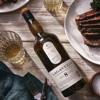 A bottle of Lagavulin 8 Year Old Single Malt Scotch Whisky sits on a wooden table beside a glass of whisky, sliced steak, and a green napkin with cutlery, evoking rich peat smoke and hearty smoked meat flavors.