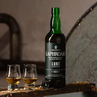 A bottle of Laphroaig Lore Single Malt Scotch Whisky stands next to two filled glasses on a wooden surface, with a barrel in the background, evoking sherried sweetness and rich peat smoke.