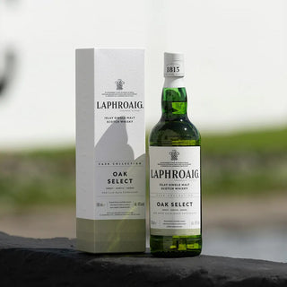 A bottle of Laphroaig Oak Select Single Malt Scotch Whisky stands beside its white and gold box on a dark surface, set against a blurred outdoor background that evokes its signature peat smoke aroma.