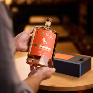 A person holds a bottle of Lark Chinotto Cask Release Tasmanian Single Malt Whisky with an orange label and bird logo over a wooden table, with its black and orange box in the background, evoking citrus and spiced herb notes.