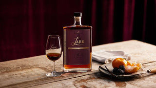 A bottle of Lark whiskey stands on a rustic wooden table beside a filled whiskey glass, with a plate of citrus and dark berries in the foreground against a deep burgundy curtain backdrop.