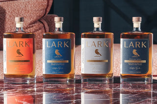 Four Lark whisky bottles in different colorways arranged in a row on a polished marble surface, with a warm textured sofa in the background.