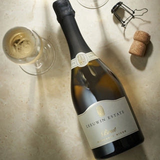 A bottle of Ieeuwin Estate Brut non-alcoholic wine with a glass of white wine, a corkscrew, and a cork on a beige stone surface.