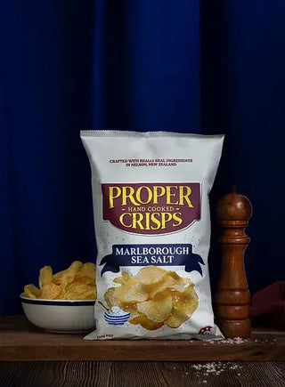 Bag of Proper Crisps Marlborough Sea Salt with chips and a bowl on a wooden surface against a blue curtain.