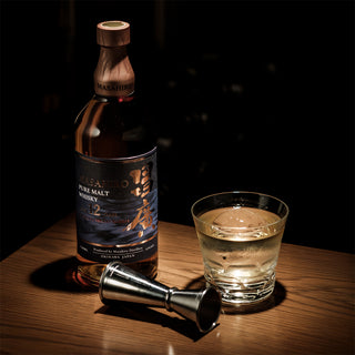 A bottle of Masahiro Pure Malt 12 Year Old Sherry Cask Japanese Whisky, a whisky glass with ice, and a metal jigger sit on wood against a dark backdrop, evoking notes of lingering oak and juicy fruits.