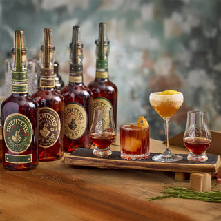 Five bottles of Michter’s US*1 Sour Mash Whiskey are arranged on a wooden surface, with three whiskey glasses and two cocktails nearby, set against a soft-focus, multicolored background with aromas of caramel and dried fruit.