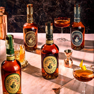 Five bottles of Michter’s US*1 Kentucky Straight Bourbon Whiskey are displayed on a marble surface with cocktail glasses, an orange twist garnish, a jigger, and a copper shaker against a dark background—evoking caramel and vanilla notes.