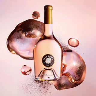 A bottle of Miraval Provence rosé wine centered against a soft pink background, surrounded by reflective rose-gold liquid droplets and powder. The bottle has a gold foil capsule and a circular black-and-gold label reading Miraval Provence.
