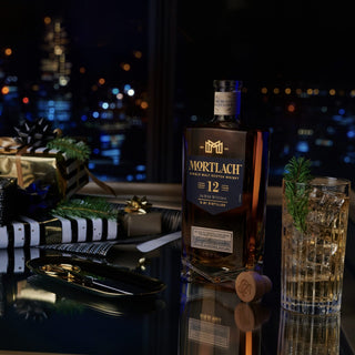 A bottle of Mortlach 12 Year Old Single Malt Scotch Whisky sits on a reflective table beside a glass with ice and pine, its rich malt aroma with hints of dried fruits, surrounded by wrapped gifts and city lights shining through the window.