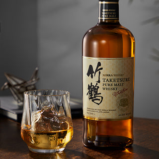 A bottle of Nikka Taketsuru Pure Malt Japanese Whisky stands beside a whisky-filled glass with ice on a wooden table, with blurred origami crane and books in the background, suggesting malt sweetness and dried fruit notes.