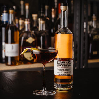A bottle of Widow Jane American Oak and Apple Wood Aged Rye Whiskey stands beside a cocktail glass with a dark drink garnished by a lemon twist, caramel toasted oak notes wafting in the air, while shelves of bottles blur in the background.