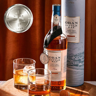 A bottle of Oban Little Bay Single Malt Scotch Whisky, silver medal, two whisky glasses with ice, and cylindrical packaging are displayed on wood—highlighting oak spice notes—against a brown curtain backdrop.