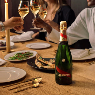 Bottle of G.H. Mumm champagne on a dinner table with people clinking glasses in the background.