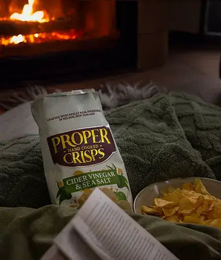 A cozy scene with a bag of Proper Crisps Cider Vinegar and Sea Salt Chips, a bowl of chips, and an open book on a blanket by a glowing fireplace.