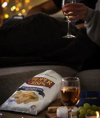 Person sitting on a couch with a bag of Proper Crisps, two glasses of wine, and snacks.
