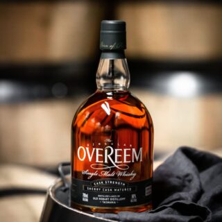 A bottle of Overeem Sherry Cask Matured – Cask Strength Tasmanian Single Malt Whisky rests on a dark surface beside a black cloth, with blurred wooden barrels in the background, evoking notes of stewed fruits and dark chocolate.