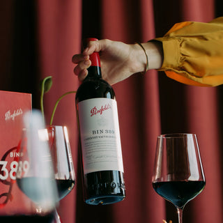 A hand in a mustard yellow sleeve holds a bottle of Penfolds Bin 389 Cabernet Sauvignon red wine against a dark red curtain background, with two wine glasses and a branded box partially visible.