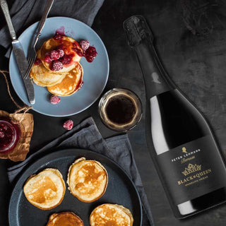 A bottle of Peter Lehmann Rose Black & Queen Sparkling Wine is styled on a dark textured table alongside pancakes on plates, a glass of red wine, and a small jar of jam.