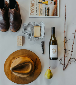 A styled flat-lay featuring a bottle of wine, a rustic hat, a pear, cheese, boots, and a magazine on a light gray surface.