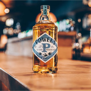 Powers Three Swallow Single Pot Still Irish Whiskey rests on a wooden bar counter, its label highlighted against blurred shelves and warm lights, inviting you to experience its rich barley flavors and subtle herbal notes.