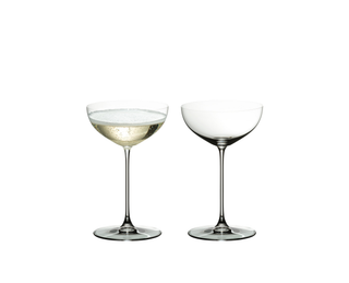 Two clear stemmed coupe glasses on a white background, one filled with a light golden beverage and one empty.