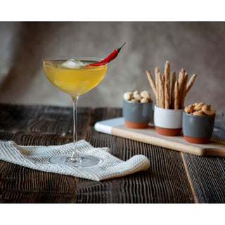A yellow cocktail in a coupe glass with a red chili pepper garnish, set on a rustic dark wood table next to small ceramic containers of snacks on a wooden board.
