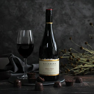 A bottle of Rockburn Central Otago Pinot Noir 2017 red wine stands on a dark slate coaster next to a filled wine glass, with chocolate-covered nuts and dried flowers arranged on a dark rustic table against a dark gray textured background.