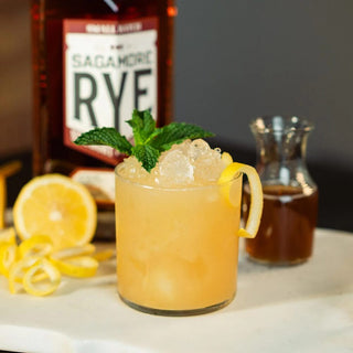 A glass of Sagamore Spirit Double Oak American Rye Whiskey, served with a lemon garnish, mint leaves, and a hint of creamy toffee.