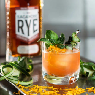 A glass of orange cocktail garnished with fresh herbs, cucumber ribbon, and orange zest sits on a reflective surface, evoking spicy rye notes, with a bottle of Sagamore Spirit Cask Strength American Rye Whiskey blurred in the background.
