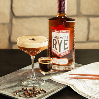 A cocktail glass with a frothy brown drink garnished with coffee beans sits beside a shot of dark liquid and Sagamore Small Batch American Rye Whiskey, evoking baking spice and orchard fruit notes on a tray with coffee beans and folded napkin.