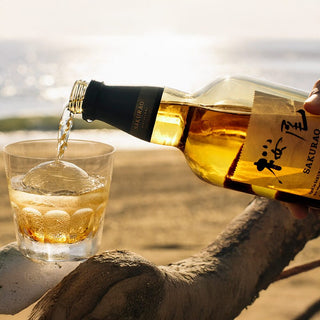 A hand pours golden Sakurao Single Malt Japanese Whisky into a glass with a round ice sphere, outdoors on a sunlit sandy beach. Aromas of ripe fruit and vanilla drift in the air, embodying relaxation and elegance.